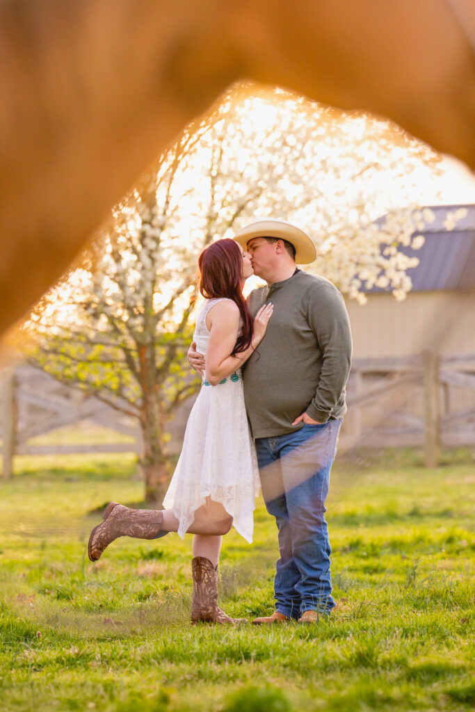 A sweet, country engagement shot at golden hour. The couple shares a kiss—she's in a white lace dress and cowboy boots, lifting one foot while wrapped up in her partner’s arms. He’s rocking jeans, a green shirt, and a cowboy hat. The shot is framed through the legs of a horse, with blooming trees and a barn adding to that rustic, heartfelt vibe