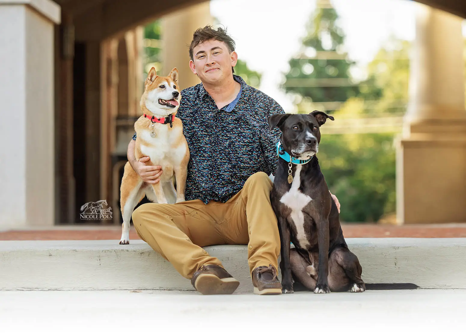 Owner Tizzy sitting on a step under a covered walkway with his pit mix and Shiba Inu leaning into him during a candid dog photography session